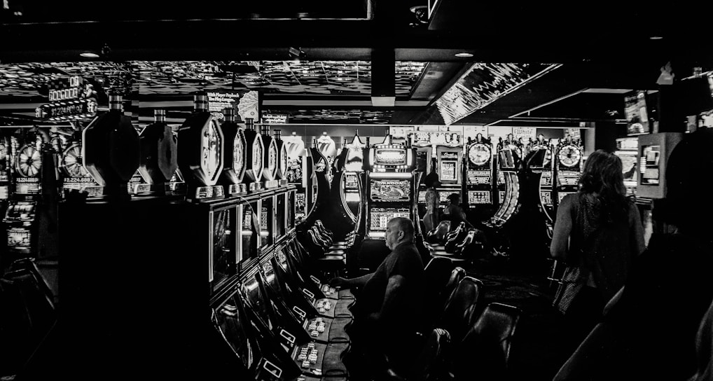 A dimly lit casino filled with rows of slot machines, some occupied by people. The machines' lights contrast with the dark surroundings, creating a vibrant yet subdued atmosphere.