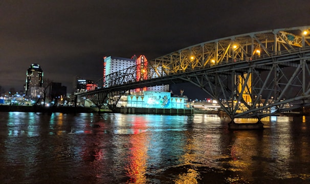 A brightly lit casino with neon signs and red lights is reflected in a river. A bridge stretches across the water, with an urban skyline visible in the background against an overcast night sky.