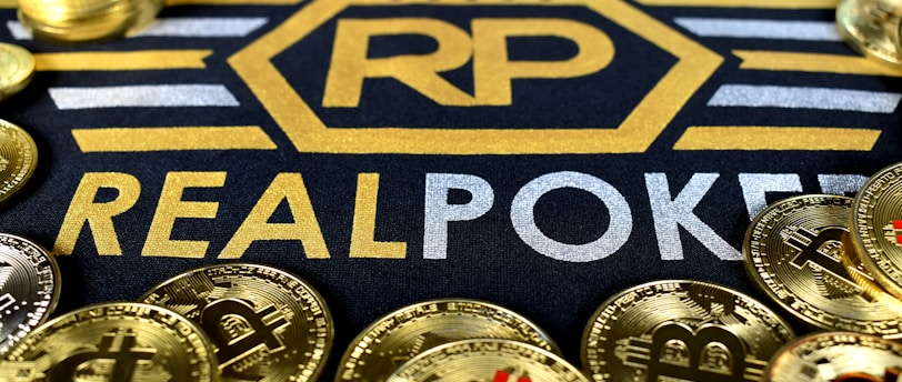 Stacks of poker chips in various colors are arranged in the background, while in the foreground, there are numerous golden coins featuring the Bitcoin symbol. The text 'REALPOKER' is prominently displayed in gold and silver lettering on a dark background.