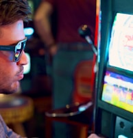 A man wearing sunglasses is intently focused on a slot machine, with colorful lights and graphics illuminating the screen. The scene is set indoors, likely in a casino or gaming area. His attire includes a patterned shirt and a blazer.