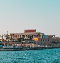 A seaside view of a large building labeled as a casino, situated on a rocky shore. The building is surrounded by palm trees and other smaller structures. The calm sea water reflects the sky, and there are several closed umbrellas lined along the waterfront.