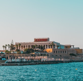 A seaside view of a large building labeled as a casino, situated on a rocky shore. The building is surrounded by palm trees and other smaller structures. The calm sea water reflects the sky, and there are several closed umbrellas lined along the waterfront.