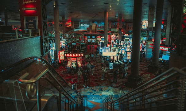 A bustling casino floor filled with groups of slot machines and people walking around. The atmosphere is vibrant with neon lights and illuminated signs. The interior has a ceiling supported by large pillars and there are stairs leading down to the gaming area.
